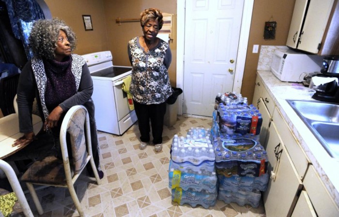 In this Thursday, Jan. 28, 2016 photo, Anna Rushing, left, 73, and her sister, Nancy Watson, right, 72, both of Flint, talk about their itchy, red and dry skin symptoms they believe they received from the lead in Flint water as they talk near cases of bottled water in the kitchen. (Todd McInturf/The Detroit News via AP)